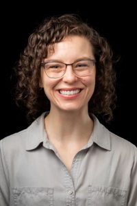Female with brown curly hair, dark eyes, wearing glasses and a light gray button down shirt.