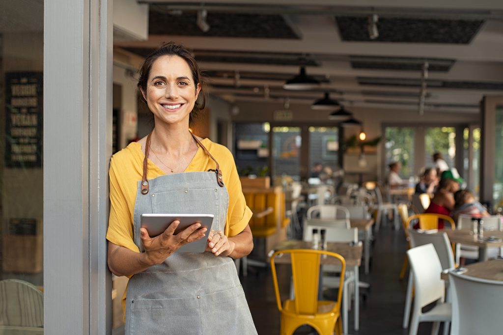 Small business owner at entrance looking at camera Portrait of happy woman standing at doorway of her store holding digital tablet. Cheerful mature waitress waiting for clients at coffee shop. Successful small business owner in casual clothing and grey apron standing at entrance and looking at camera.