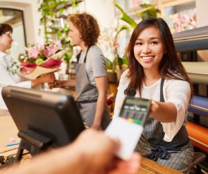 A smiling cashier holds out a credit card reader to a hand holding a credit card. They are in a flower shop.
