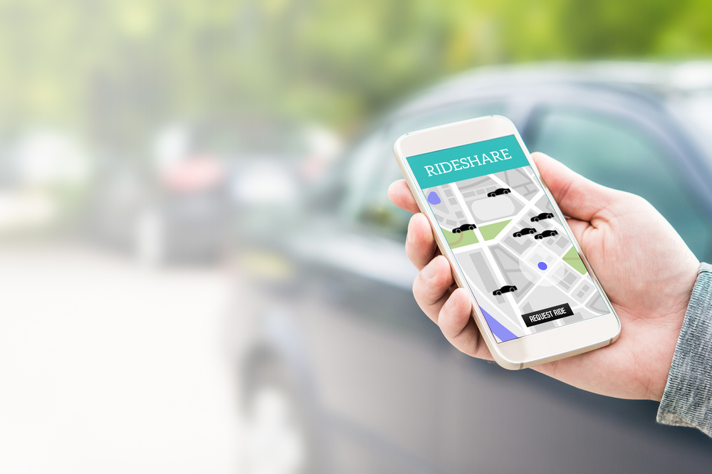 Rideshare taxi app on smartphone screen. Online ride sharing and carpool mobile application. Modern people and commuter transportation service. Man holding phone with a car in background.