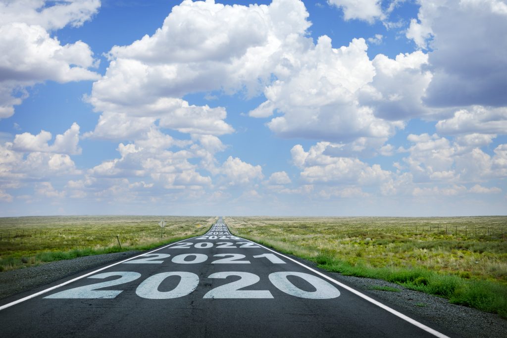 The year 2020 and subsequent years are painted on to a long straight rural highway that disappears at the horizon under a canopy of cumulus clouds.