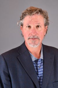 man in blue striped collared shirt with dark grey blazer. Curly hair