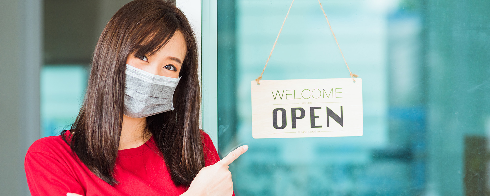 woman with mask on pointing at welcome we are open sign
