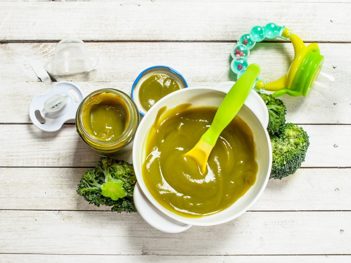 Green baby puree in a bowl with a green spoon next to a pacifier and baby toy. 