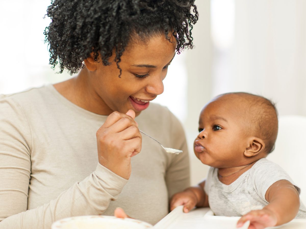A woman in beige spoon feeds an infant.