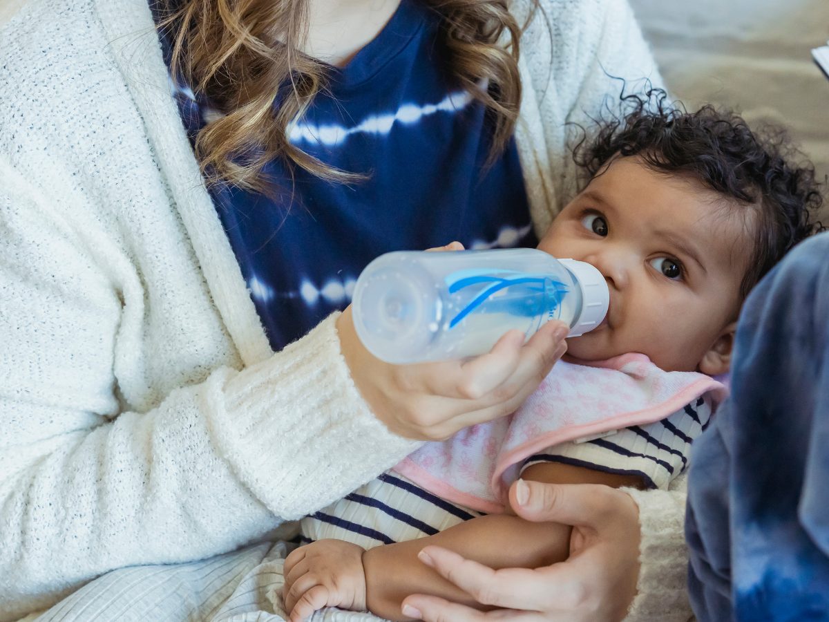 Baby drinks from bottle while gazing at camera from woman's arms.