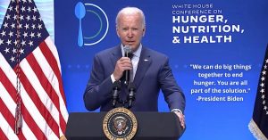 President Biden speaking at conference on hunger, nutrition and health, standing behind podium in front of blue background and American flag
