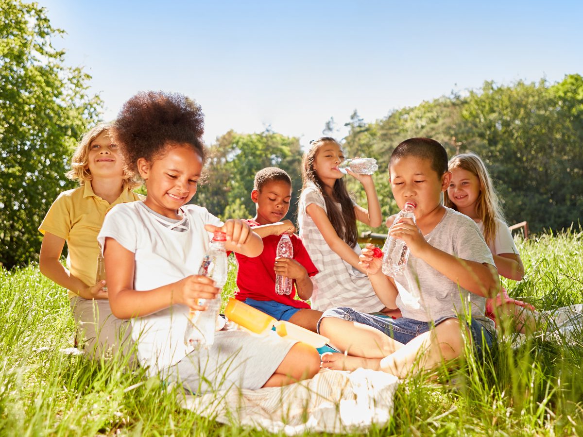 Diverse group of children sitting in the grass eating healthy snacks and drinking water.
