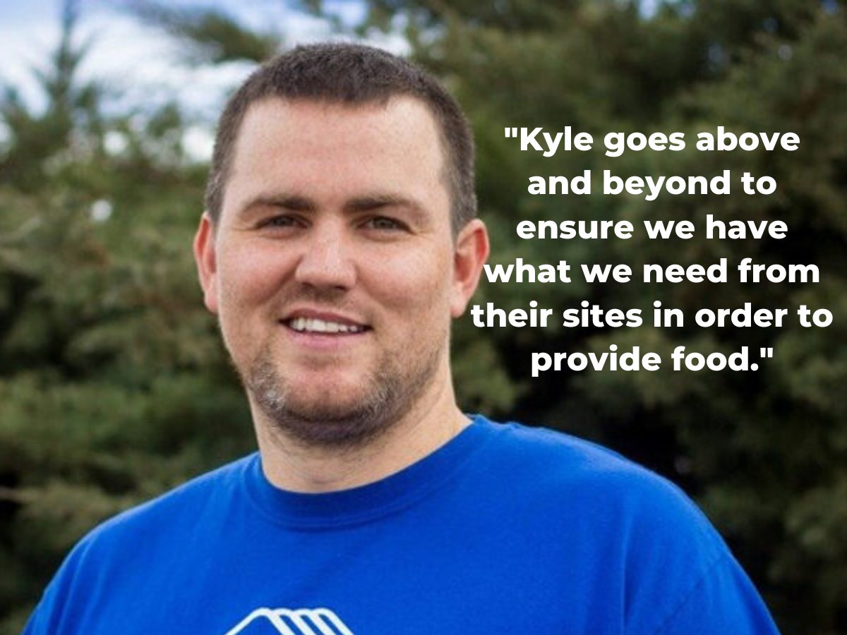 An adult man, Kyle Jackson, wearing a blue t-shirt stands in front of evergreen trees.