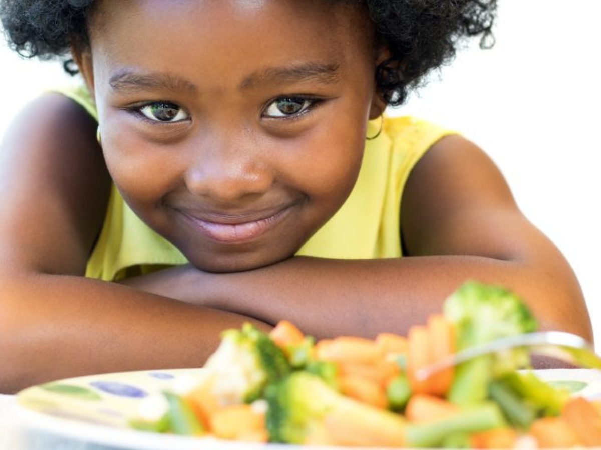 Young black girl smiling and leaning on table with a plate of orange and green veggies in front of her