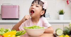 Young Asian girl eating a cherry tomato with a bowl of lettuce in front of her and other vegetables on the table