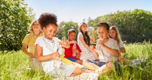 Diverse group of children sitting in the grass eating healthy snacks and drinking water.