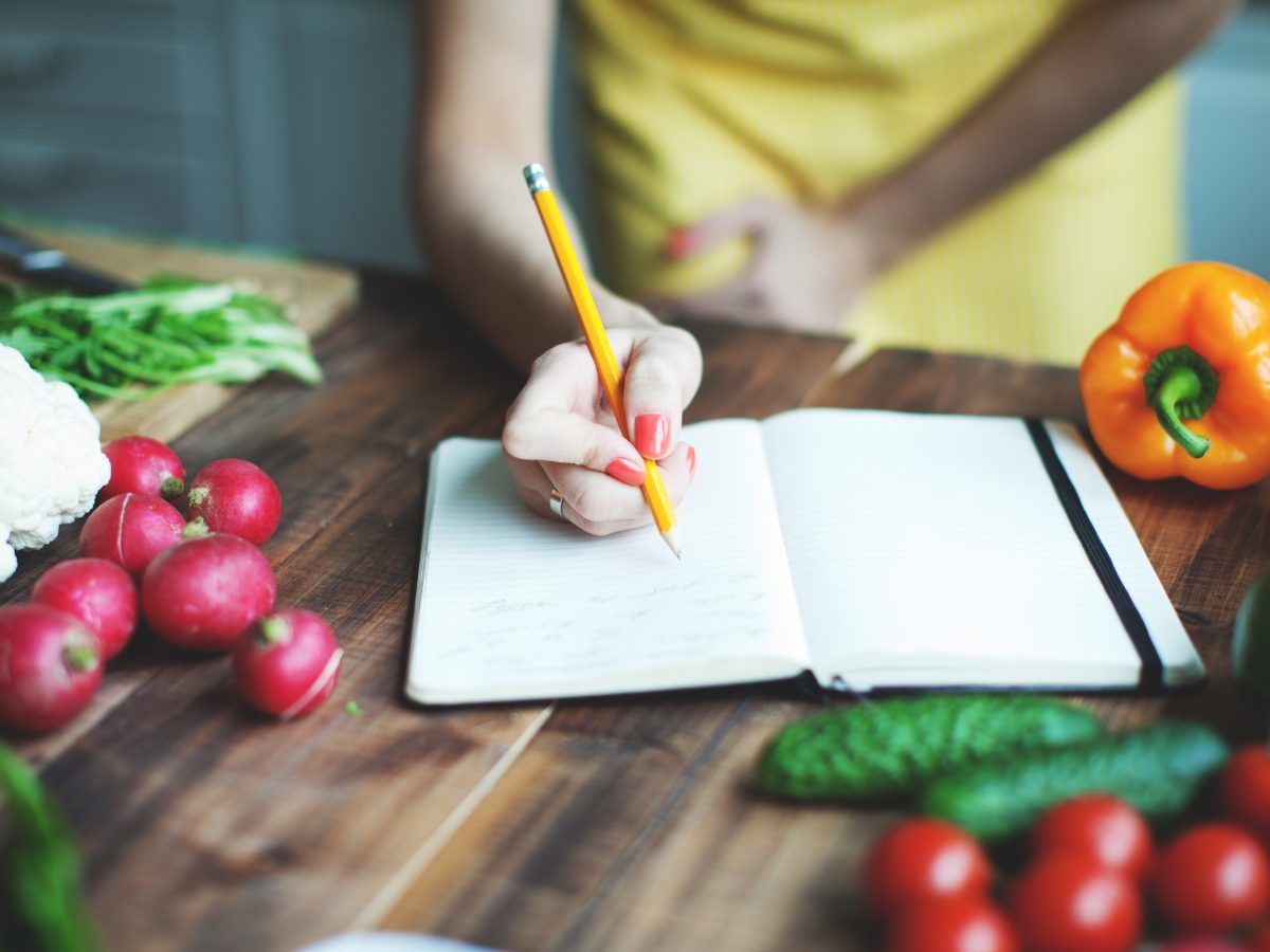 A wooden counter top holds vegetables and an open notebook. There is a woman's hand holding a pencil over the notebook.