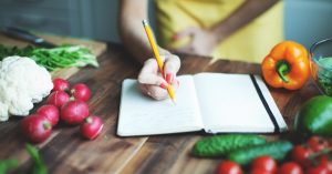 A wooden counter top holds vegetables and an open notebook. There is a woman's hand holding a pencil over the notebook.