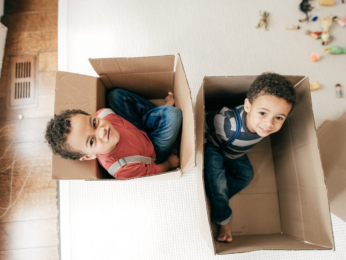 Two young boys sit side by side in two open cardboard boxes on the floor. They are smiling, playing a CACFP relay game.