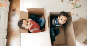 Two young boys sit side by side in two open cardboard boxes on the floor. They are smiling, playing a CACFP relay game.
