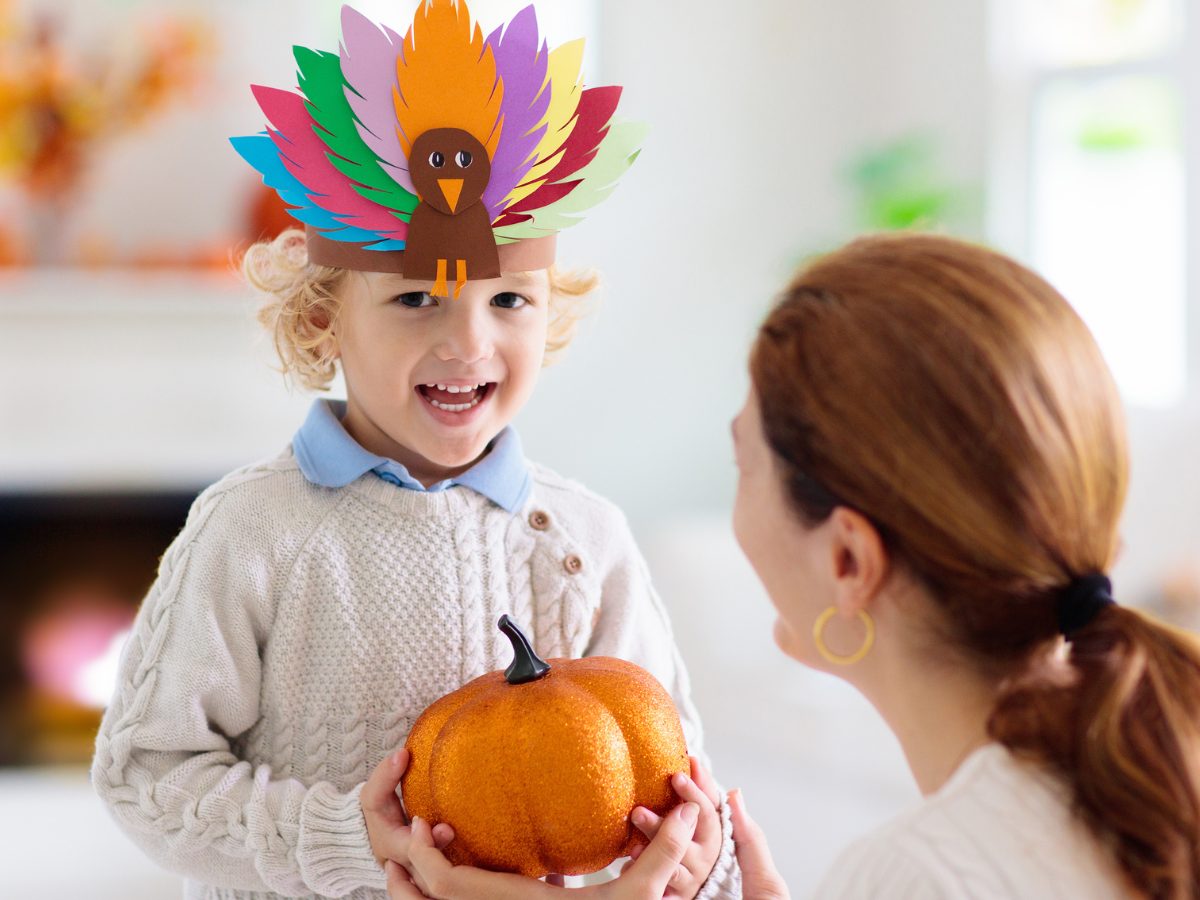 A child wears a headband with an arts and crafts construction paper turkey on it. In front of him, a woman is kneeling down and handing him a sparkly orange pumpkin.