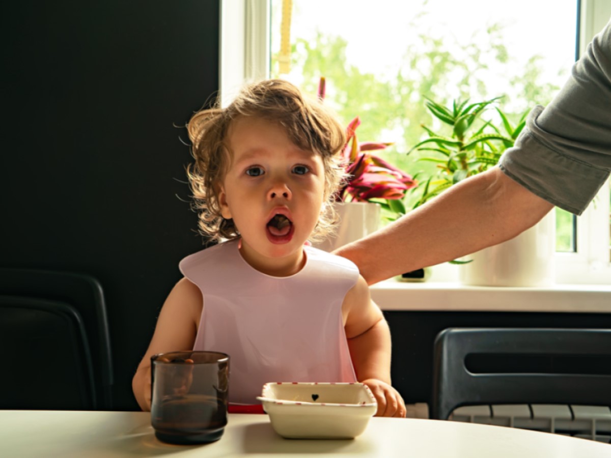 A small blond girl coughing with her mouth open while her father pats her on the back to dislodge any food that may have gone down her throat.