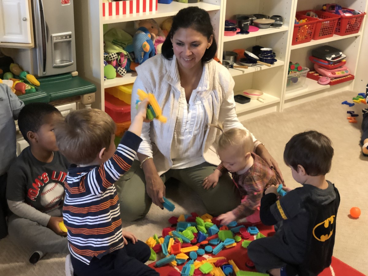 Female Family Child Care Home provider sits in a circle with young children playing with toy building blocks