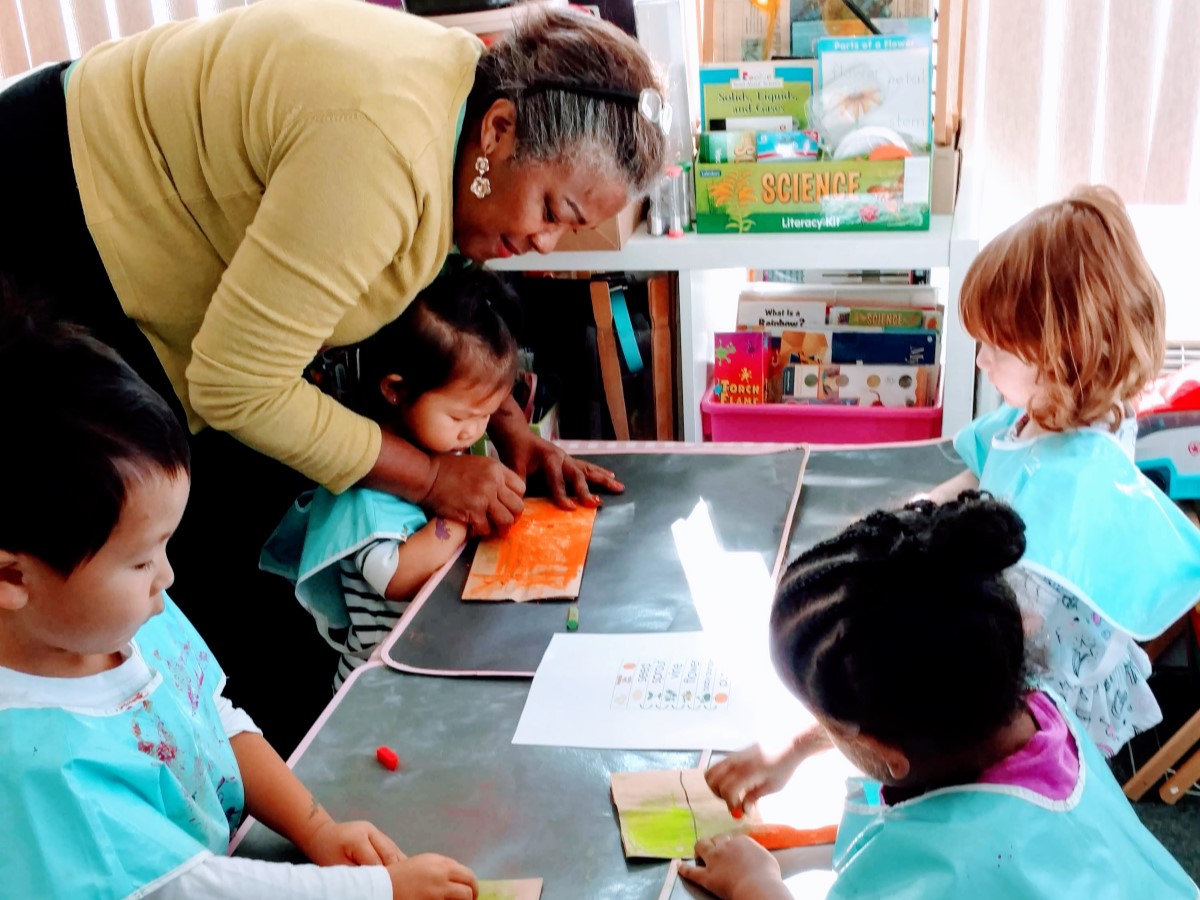 Female child care provider leaning over a table helping children with craft.