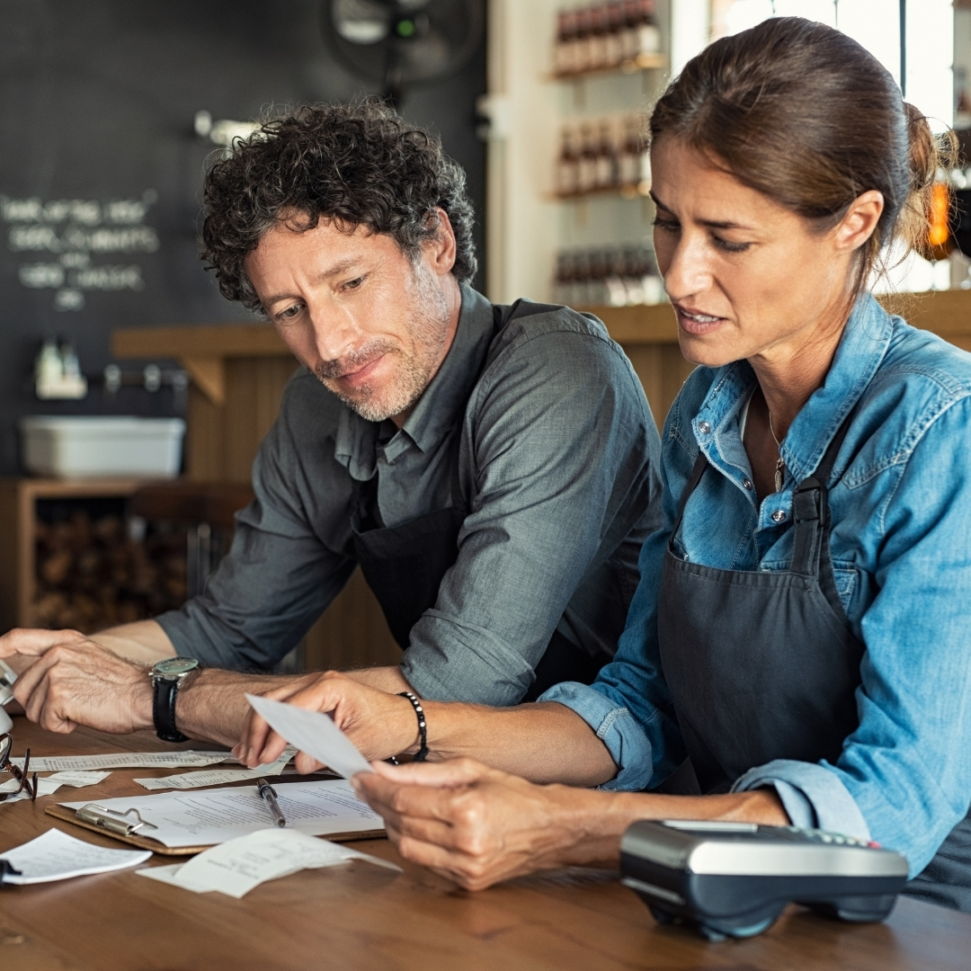 A white man and woman sitting at a table in a small cafe shot looking concerned over a pile of receipts in front of them.