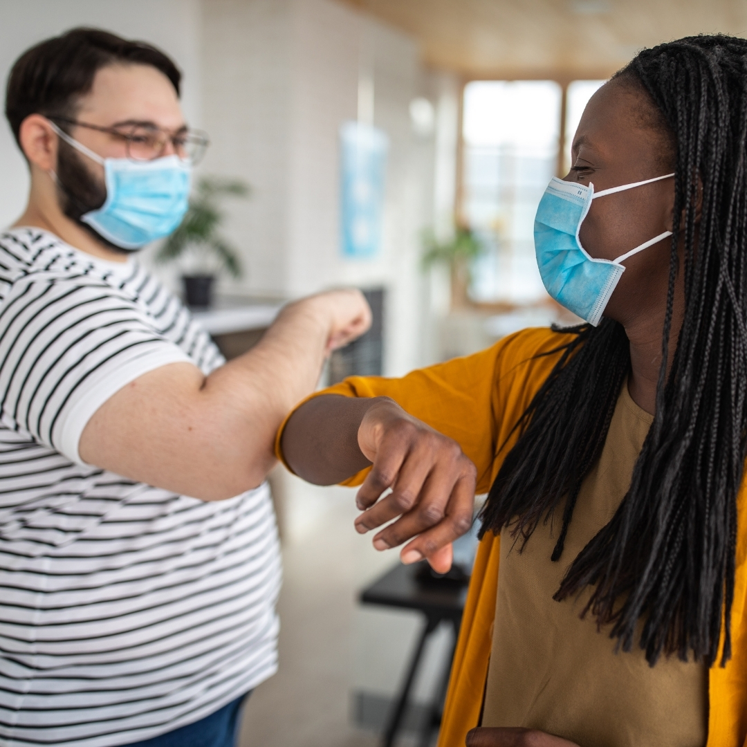 A white man wearing a face mask elbow bumping a black woman wearing a face mask.