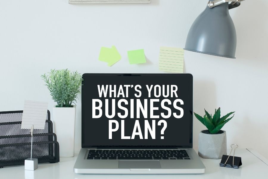 An open laptop with the phrase, "What's Your Business Plan" displayed on the screen sitting atop a white desk with a variety of office related supplies.