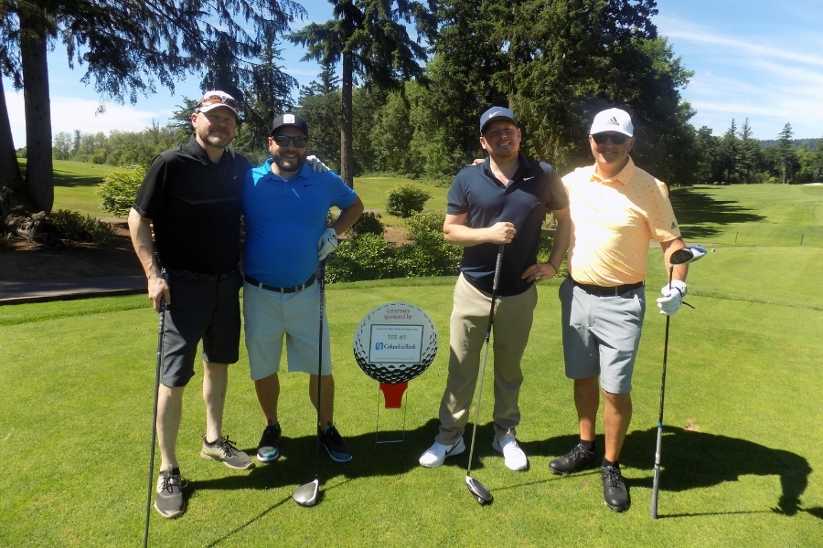 Picture of four white men in golf attire and baseball hats holding driving clubs next while standing beside a sign that reads, "Tee #1 Generously sponsored by Columbia Bank."