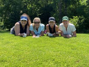 Four white women pose in a row on green grass. They are lying on their stomachs, using their elbows to support them and they are smiling at the camera.