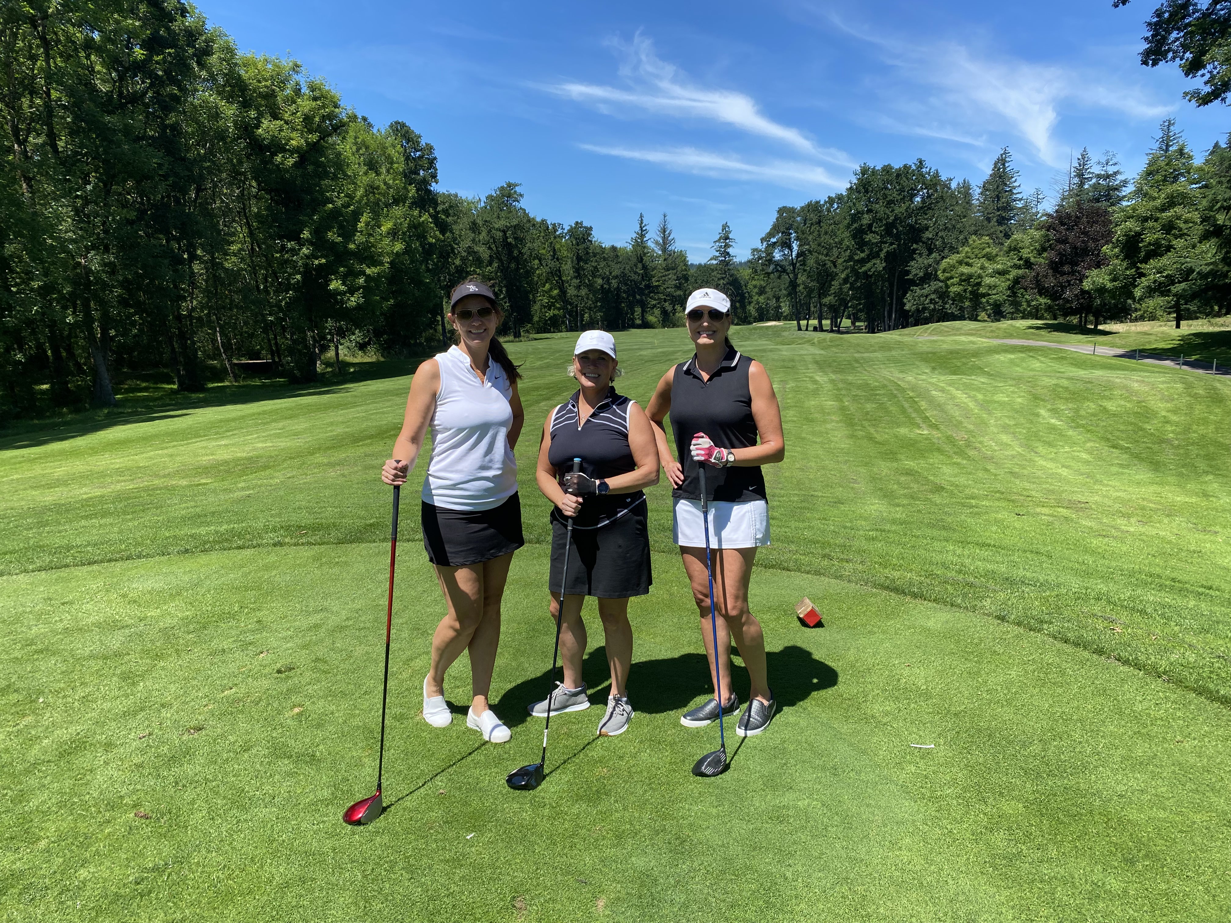 Three women golfers pose on the tee box with their clubs, smiling.