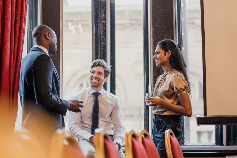 A profile view of a black man standing in a suit talking to a white man in a shirt and tie who is sitting down and smiling and a brown skinned woman who is standing and smiling while holding a glass of water..