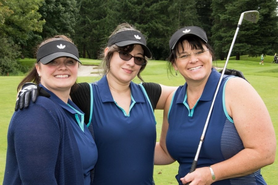 SSS_Ladies_Golf_Tournament Three women in matching blue golf shirts with black visors with their arms around each other facing the viewer smiling with the far right woman holding a golf iron resting on her shoulder.