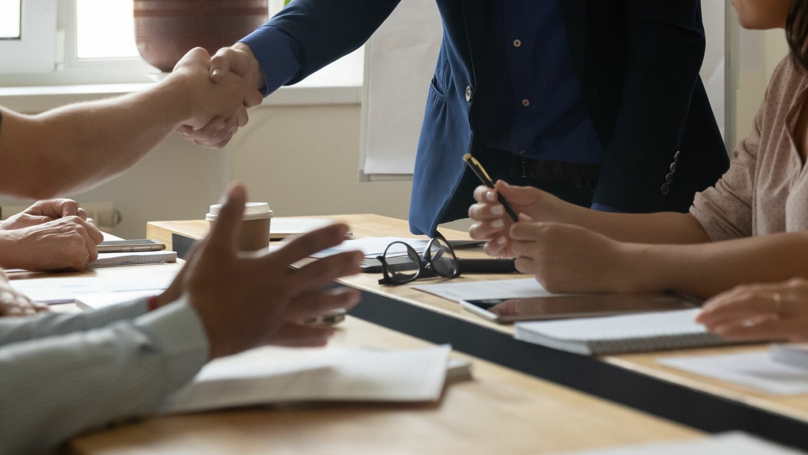 Close up photo of multiple people of different genders and ethnicities at a tableduring a meeting. Focus is on their hands and forearms. One pair of individuals is standing and shaking hands.