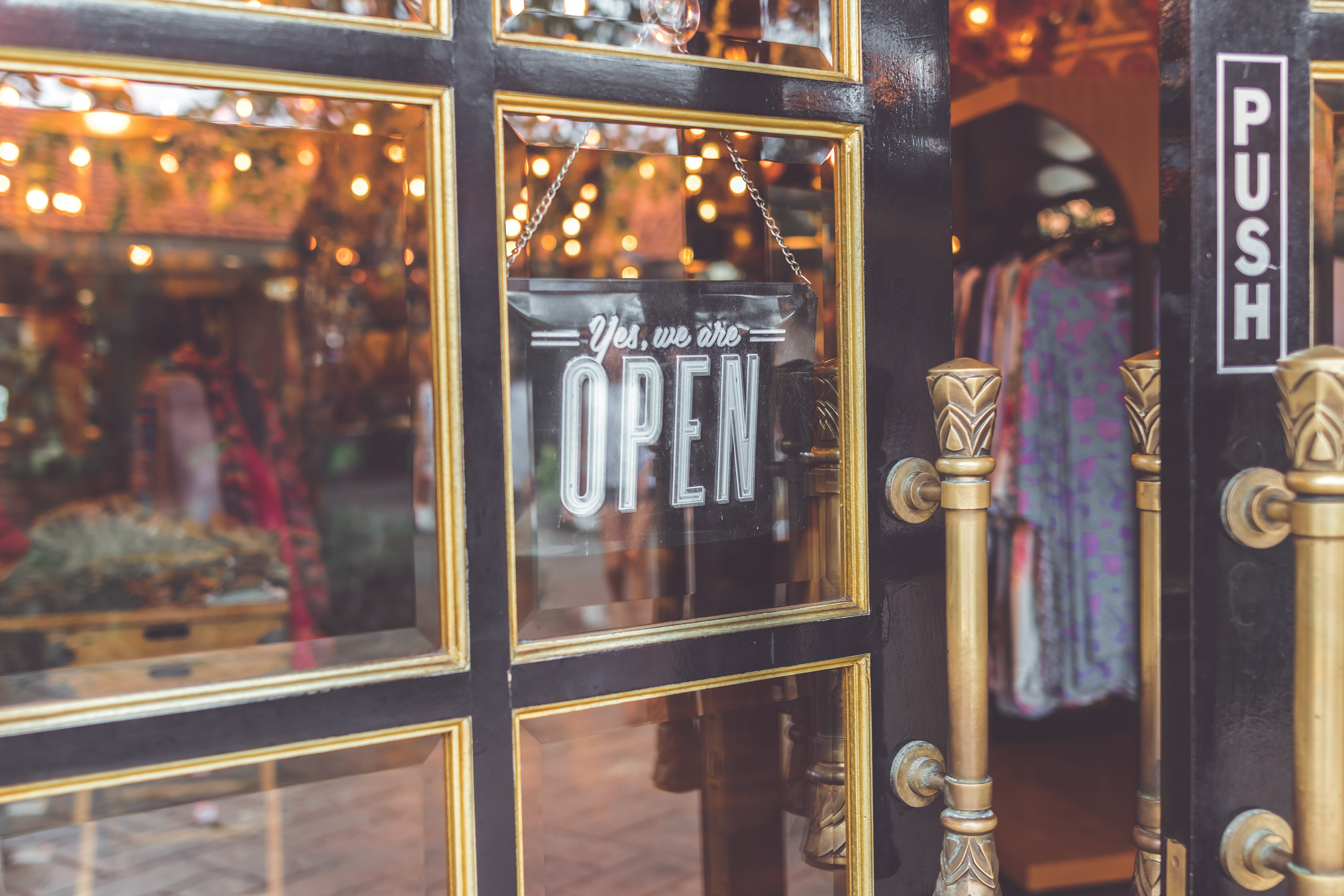 A fashion clothing store with the door propped open and a sign in the door's window saying "Come in we're open."