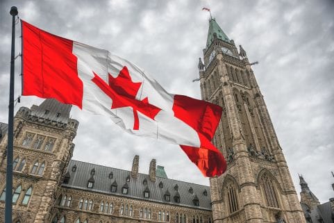 A photo looking up at the Parliament buildings in Ottawa. There are overcast skies above and a Canadian flag is waving across the front of the building.
