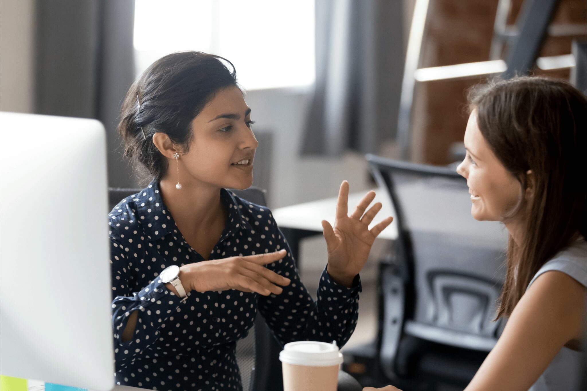 Two women in an office setting, engaged in a conversation, smiling at each other.