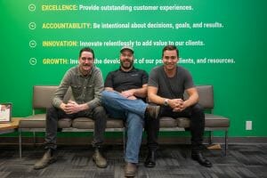 Three young men sitting side-by-side on a couch, facing the camera and smiling. There is a green background behind them with some of the words from their company's value/purpose statements on the wall.