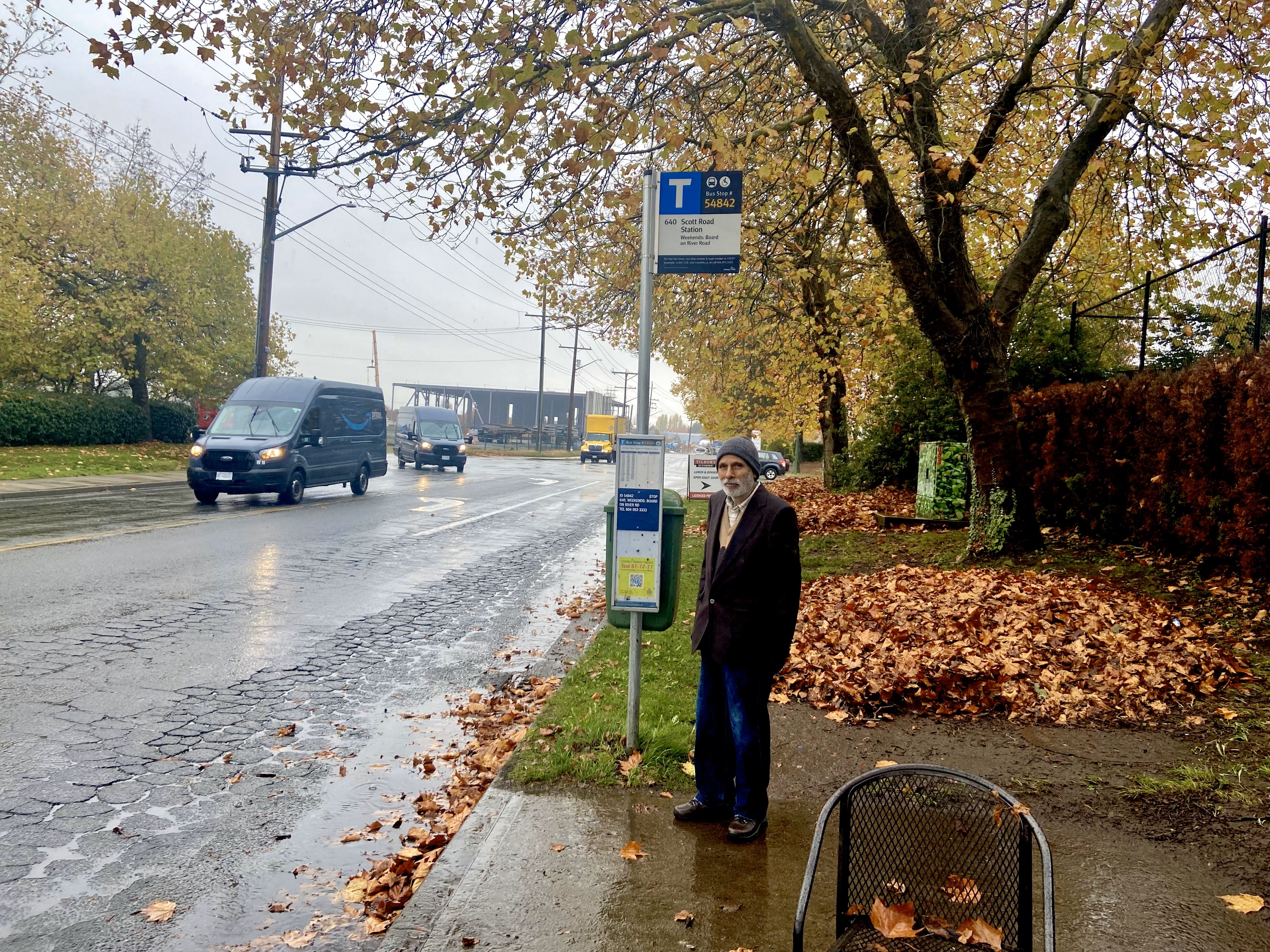 A middle aged South Asian man wearing a jacket and hat stands at a bus stop. The sky is grey and raining. It is Autumn with leaves on the ground. Two Amazon delivery trucks are driving past.
