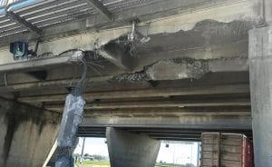 Image shows the underside of the Highway 17A overpass of the southbound side of Highway 99. Concrete and metal is torn away and hanging off the underside of the overpass. The oversized commercial container truck which struck the overpass is stopped and remains visible in the far right corner of the photo.