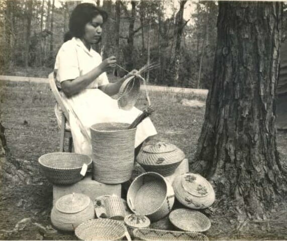 Coushatta Image 15 Ena Mae Sickey poses for a photo sewing a pine needle basket in front of a large collection of pine needle baskets, c. 1960s.
Images may not be reproduced or used in any way without the express written permission of the Coushatta Tribe of Louisiana Tribal Archives.