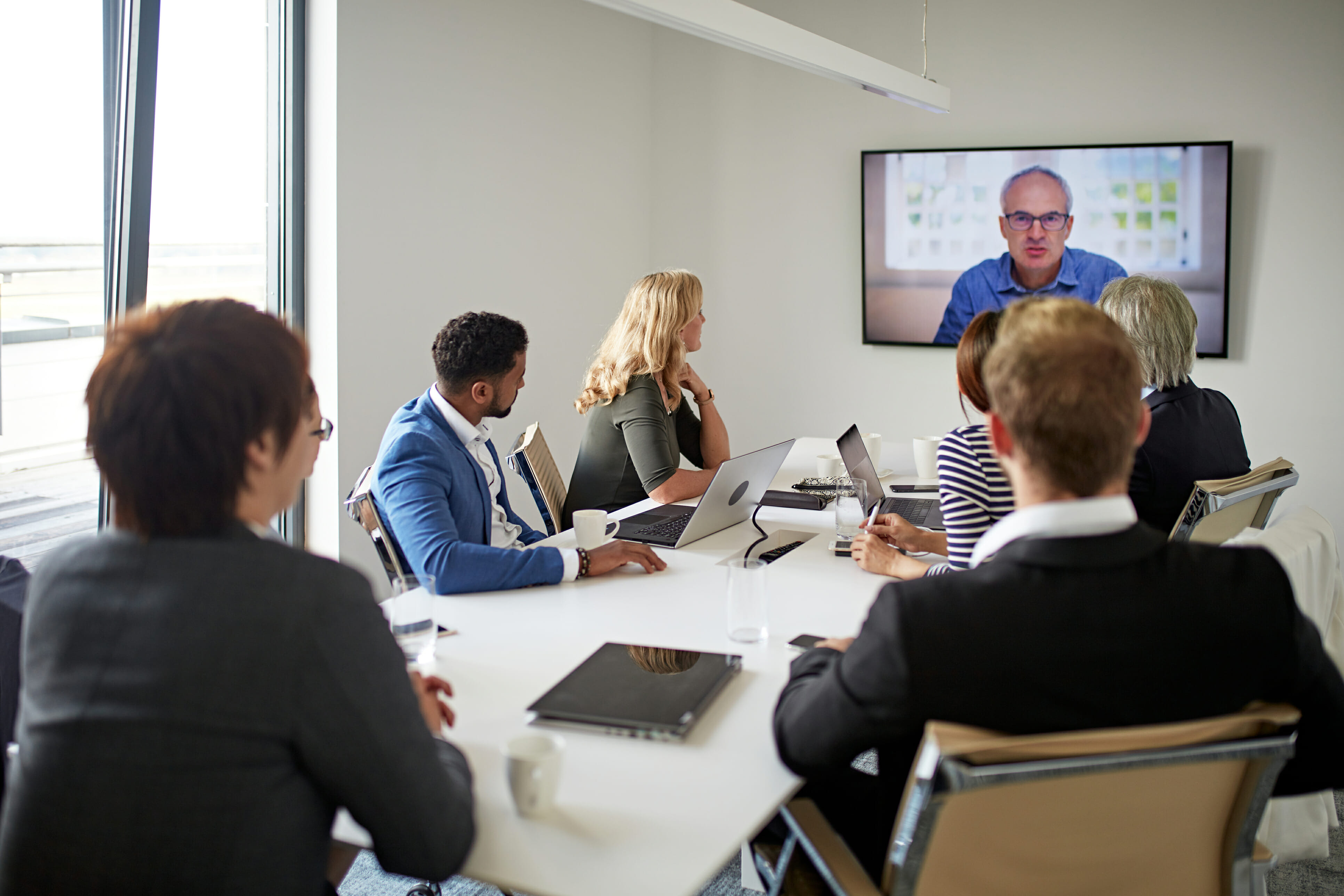 Diverse group of businesspeople sitting in modern board room conversing with male CEO on big screen television.