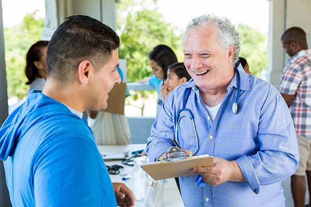 Senior Caucasian male doctor talks with mid adult Hispanic male patient at outdoor health fair. The doctor is collecting information from the patient.