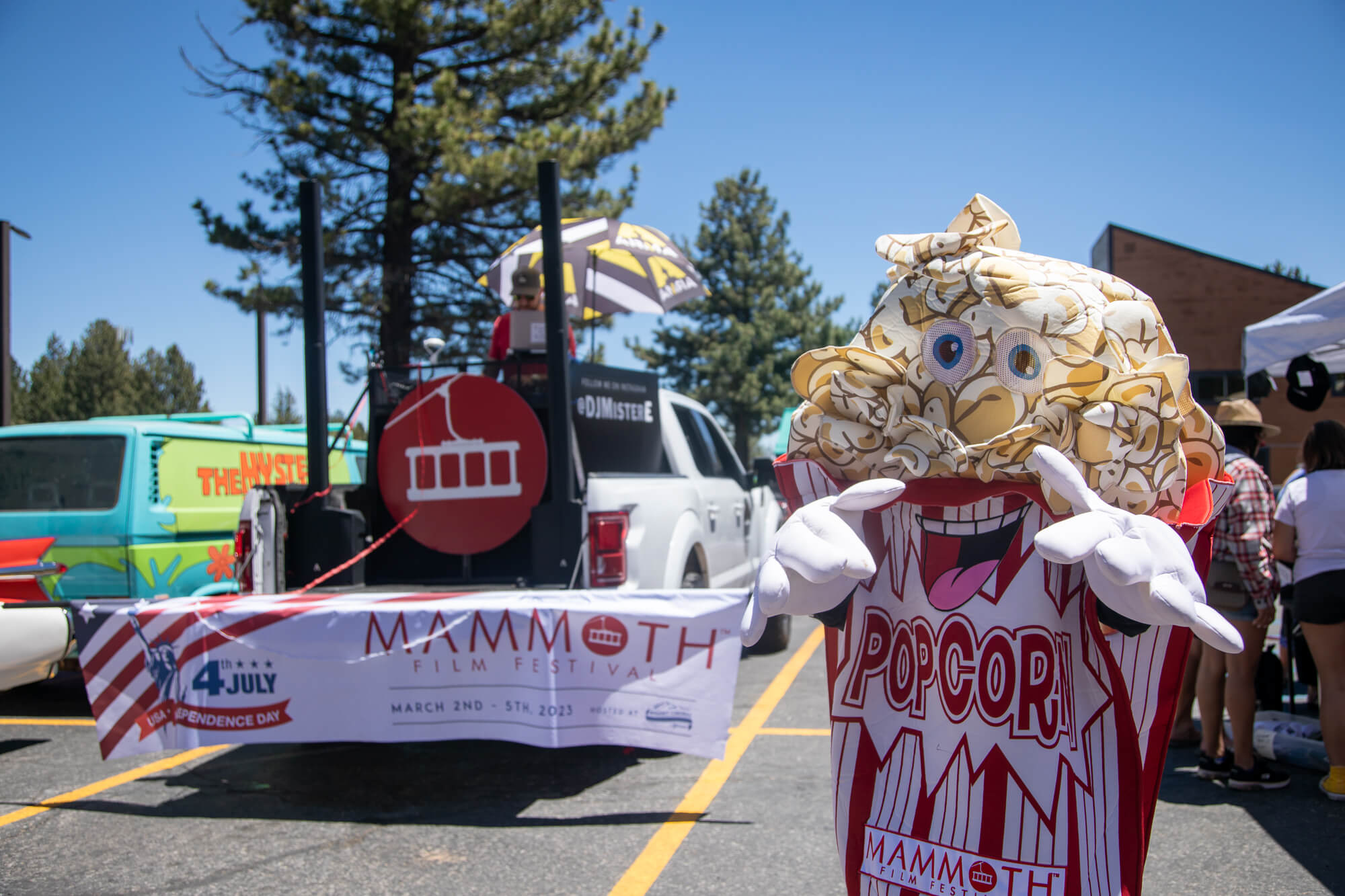 A person dressed as popcorn smiles at the camera in front of the Mammoth Film Festival float in the Mammoth Lakes 4th of July parade