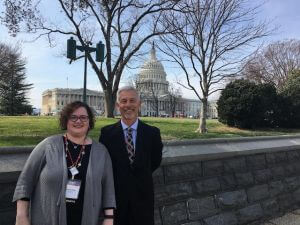 Rachel Fink and Brad Erickson standing in front of the Capitol Building. Rachel is a light skinned woman with short dark curly hair and glasses. She is wearing a grey cardigan, black dress, neclaces and a name tag on a red chord around her neck. Brad is a white man with short grey hair and a close shaved beard. He is wearing a black suit, blue button up, and a black plaid tie.