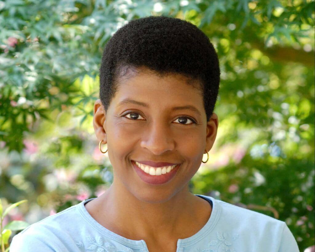 Headshot of Velina Brown. She is a black woman with short cropped hair. She is wearing a light blue shirt and standing in front of green foliage. She is smiling with her teeth showing.