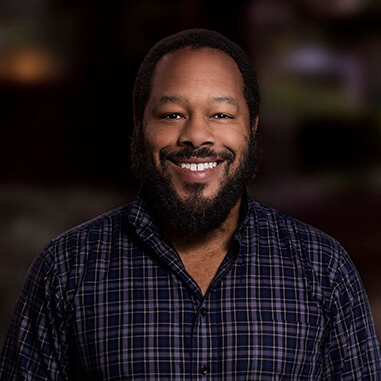 Michael Wayne Rice wearing a blue plaid shirt. He is a black man with short black hair and a beard and goatee. he is smiling with his teeth showing.