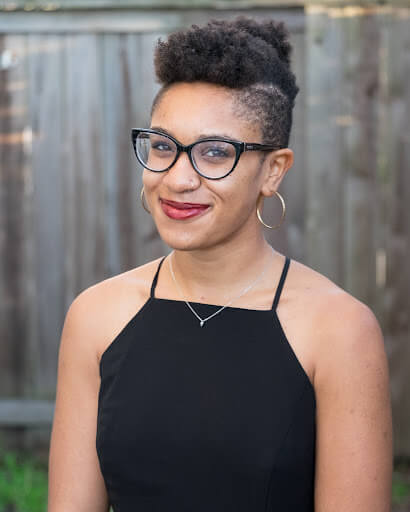 Leigh Rondon-Davis is wearing a black tank top, gold hoop earrings, and large black rounded glasses. They are a light skinned black person with a haircut that is longer on the top and shorter on the sides. They are smiling and wearing orange red lipstick. 