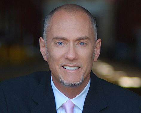 Headshot of Alan Brown in a black suit, white shirt, and pink tie. He is a white man with blue eyes. He has short shaved grey hair, and a grey goatee. He is smiling showing his teeth.