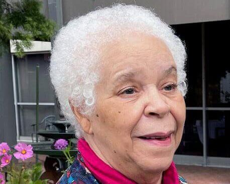 Anne Smith in a colorful sweater smiles, looking off to the right of the camera. She is a Black woman with short white hair.