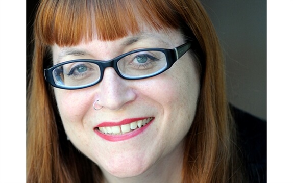 Headshot of Melissa Hillman. A light skinned woman with blue eyes, black oval glasses and wearing red lipstick with a nose ring. She  has red hair with bangs. She is smiling with her teeth showing. 